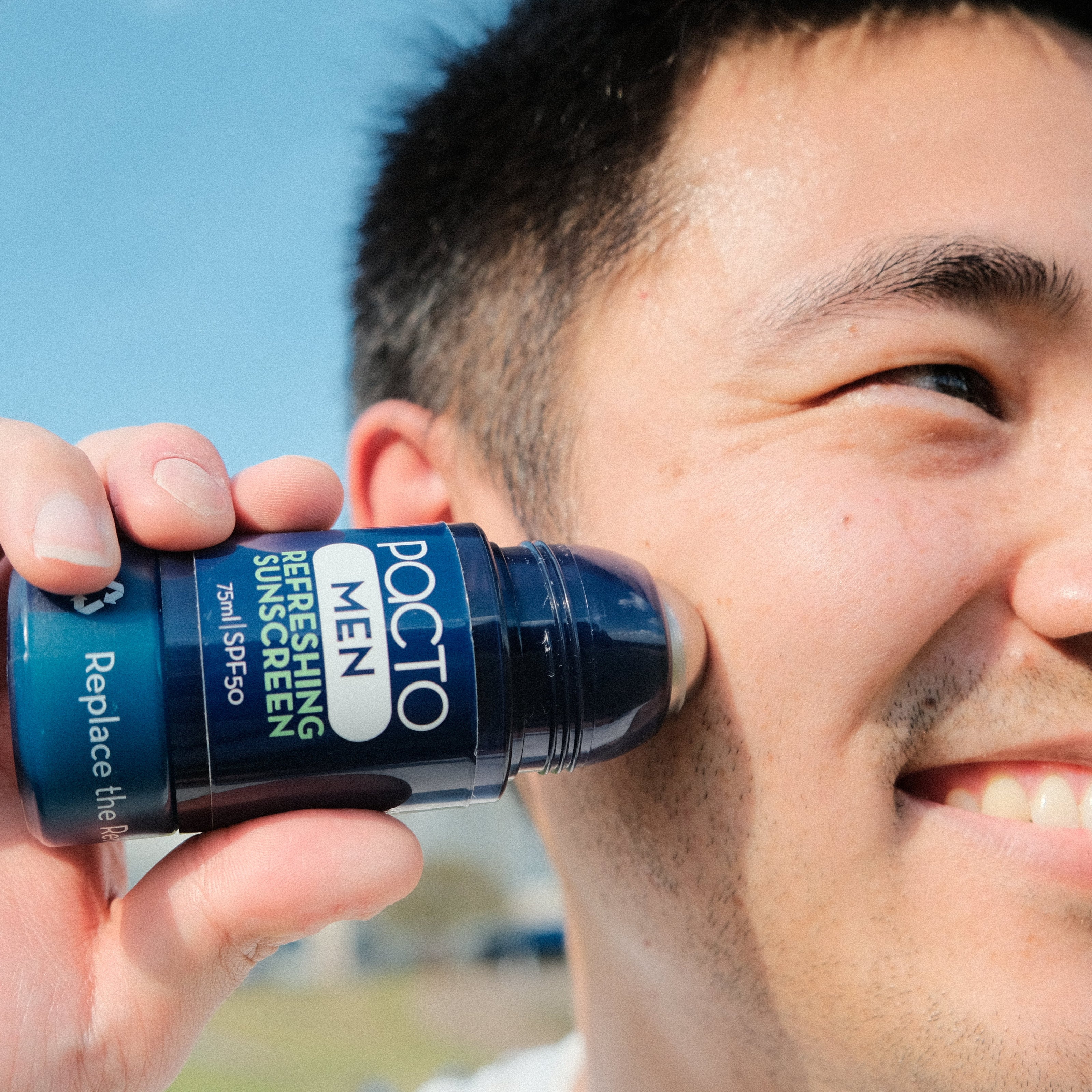 Man applying a bottle of 'Pacto Men' sunscreen to his face with a clear sky background